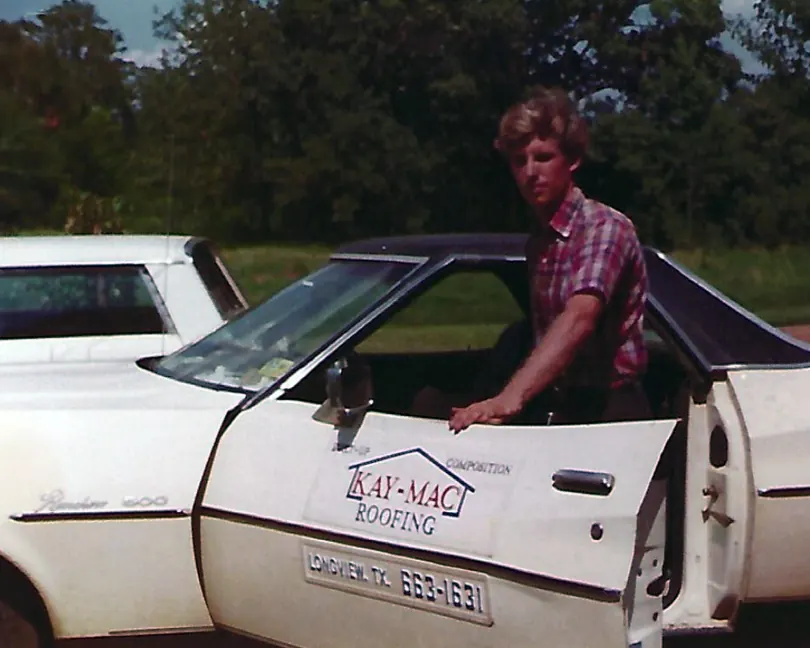 A young man standing beside a white car with KAY-MAC ROOFING, Longview TX lettered on the door