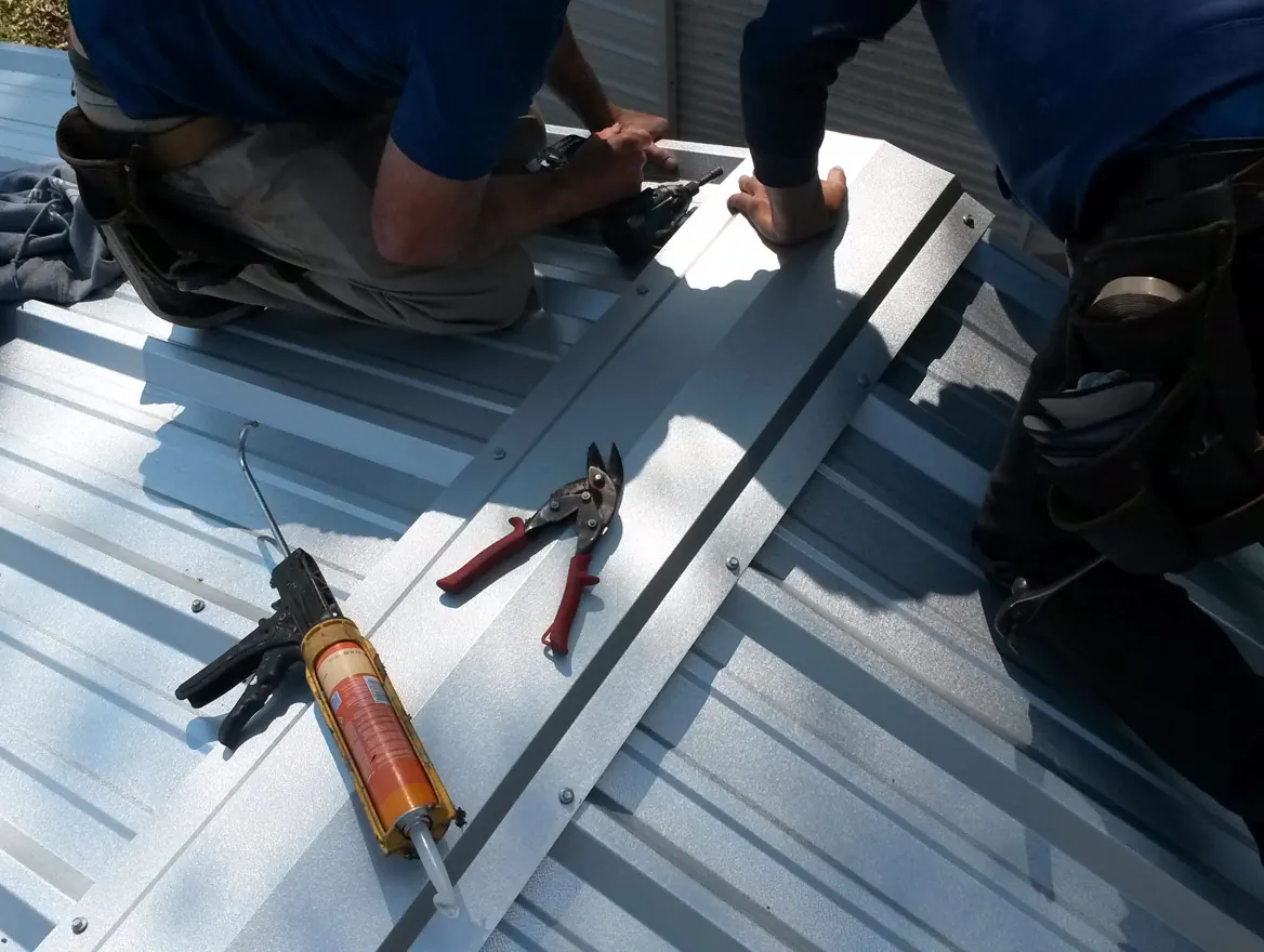 Two workers fastening a metal roof panel, with a caulk gun lying on the seam