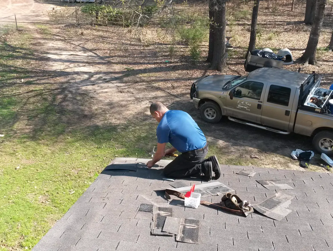 A roofer kneeling on a gray shingle roof beside his work truck, replacing damaged shingles